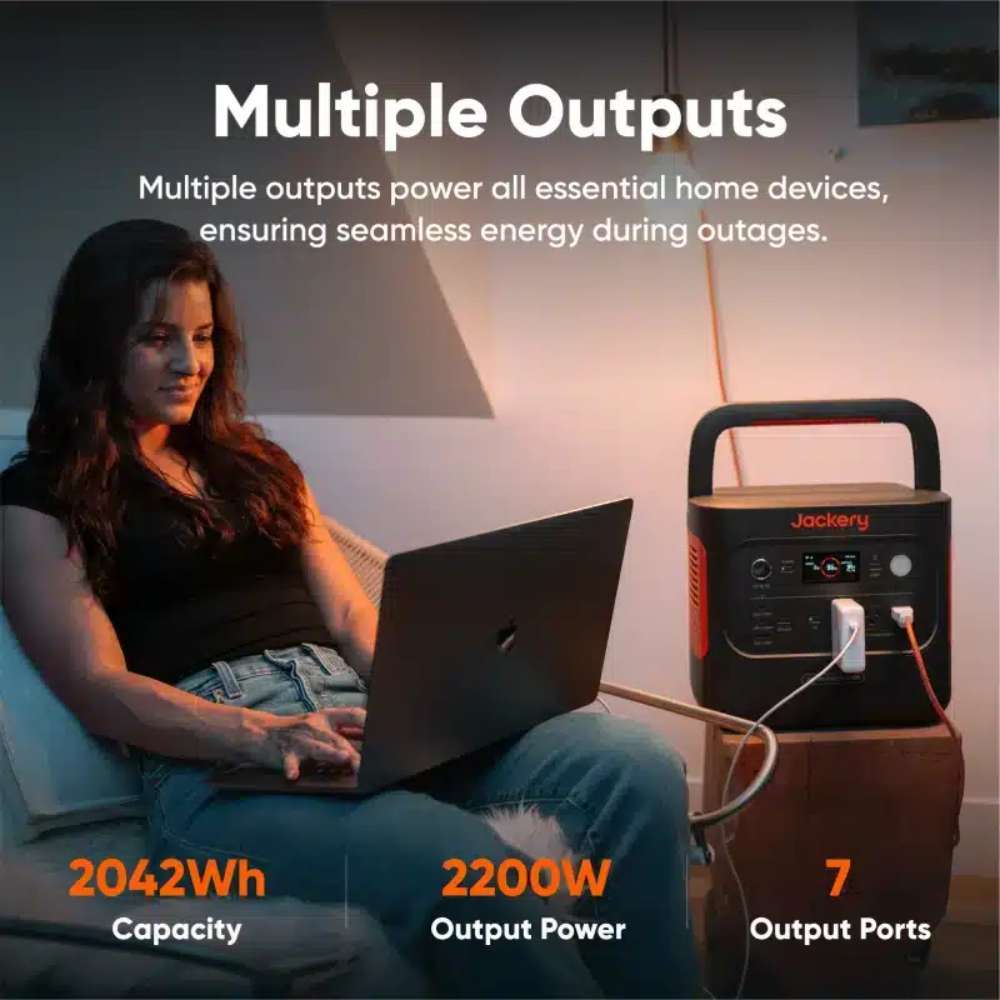 Jackery Explorer 2000 V2 Portable Power Station sits beside a woman using a laptop, showing multiple output ports for powering home devices during an outage
