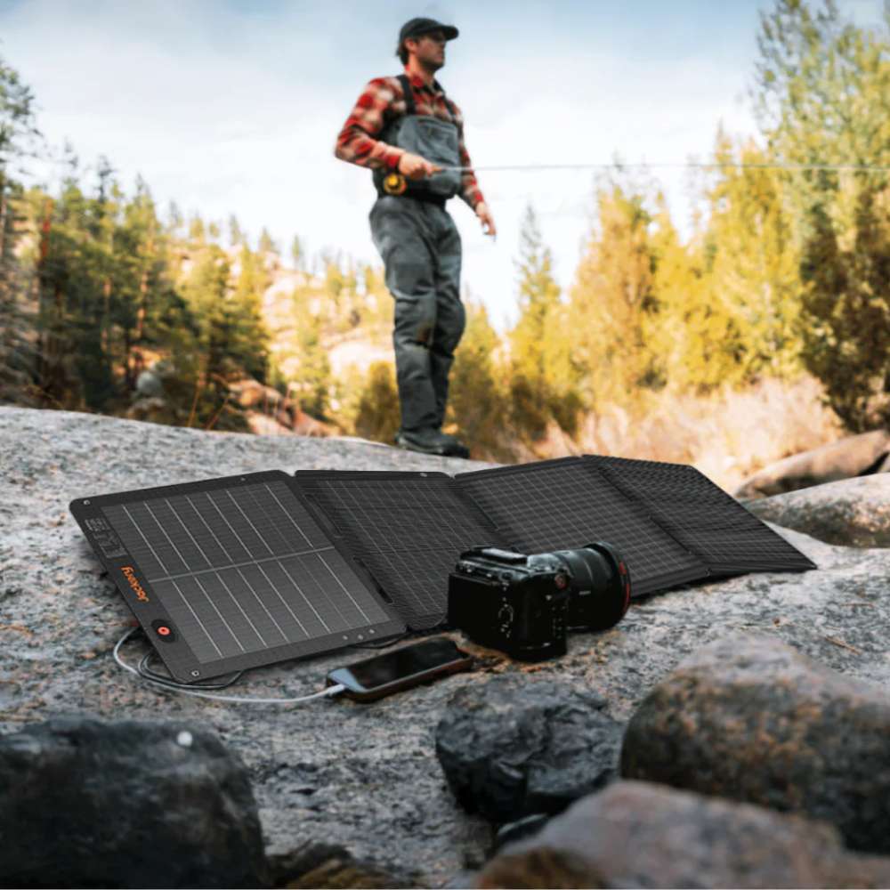 Jackery SolarSaga 100W Air Solar Panel charging a phone and camera on a riverside while a person fishes in the background