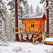 Warm wood Jumbo Grand Loft 130 SD cabin elevated on a deck amid snow-covered pine trees and falling snow.