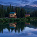 Two wooden cabins beside a still lake at dusk, with the Jumbo Grand Loft 130 SD on the left and both buildings reflected in the water.