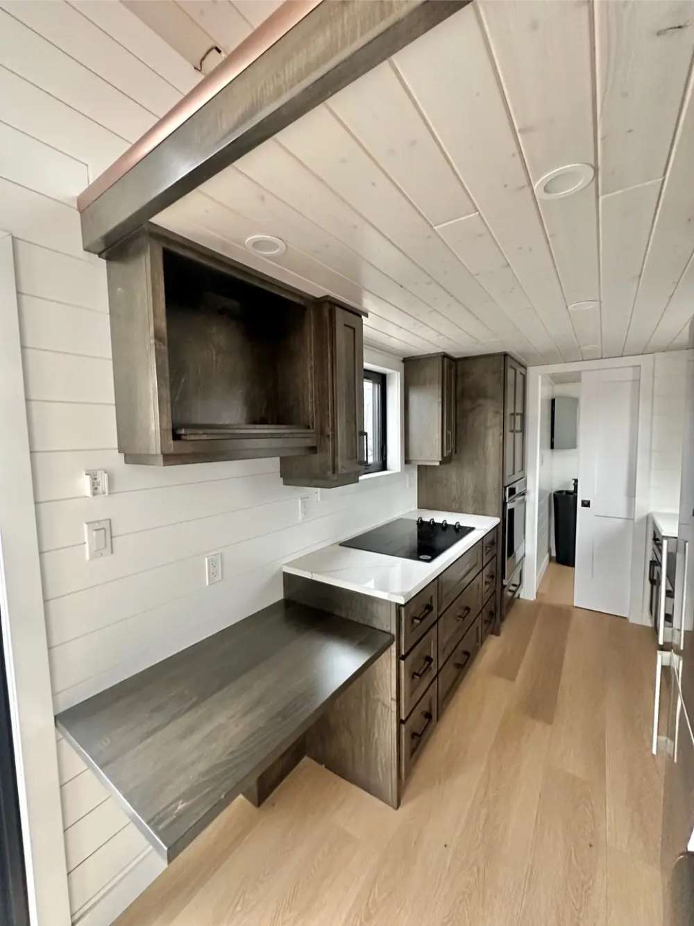 Kitchen in Sawmill Structures Wanderlove Tiny Home includes dark wood cabinets, a cooktop, and a small work desk beneath a wood beam