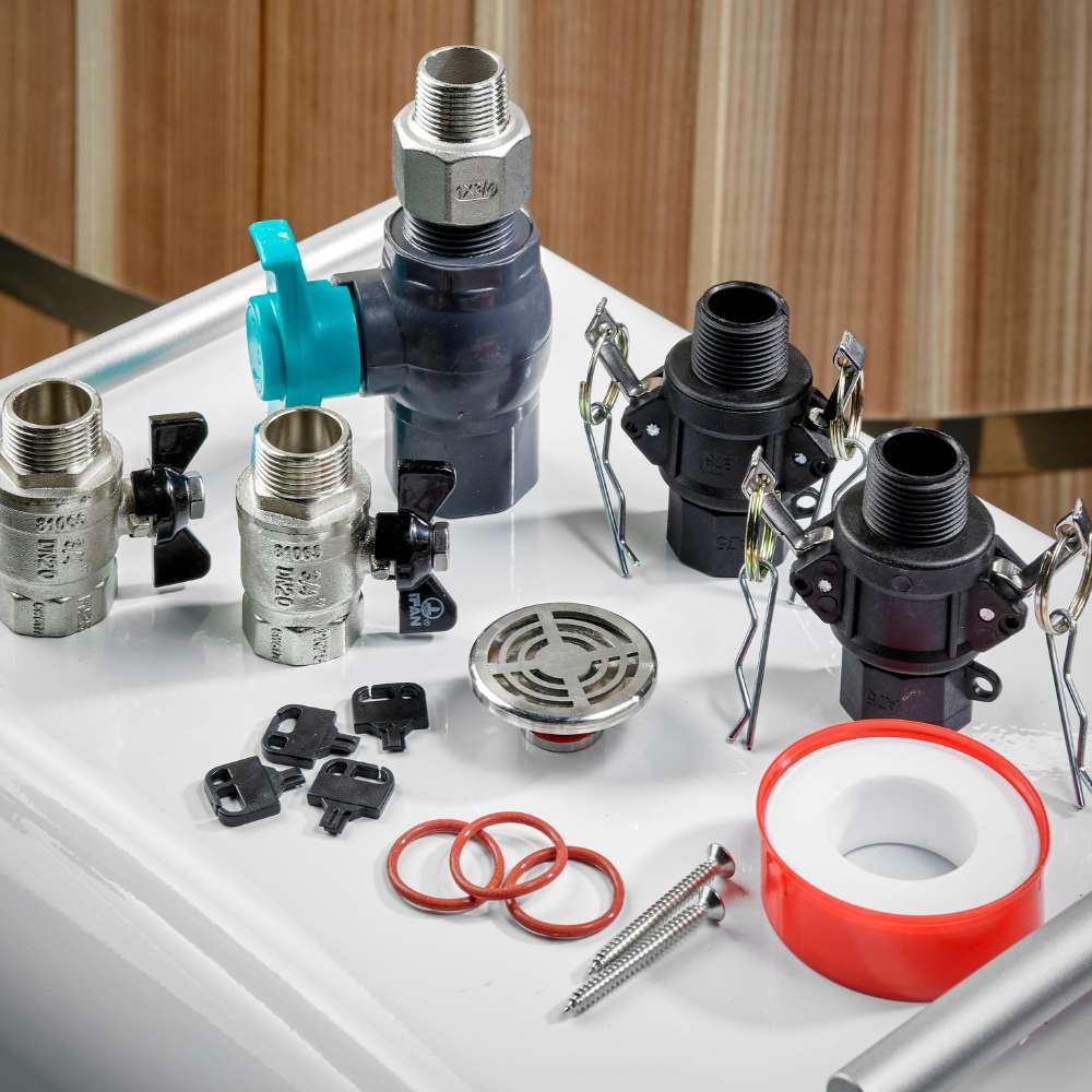 Laid out on a white surface, the valves, hose connectors, drain grate, O-rings and thread seal tape for Golden Designs Dynamic Cold Therapy PVC Barrel Cold Plunge sit in front of a cedar wall