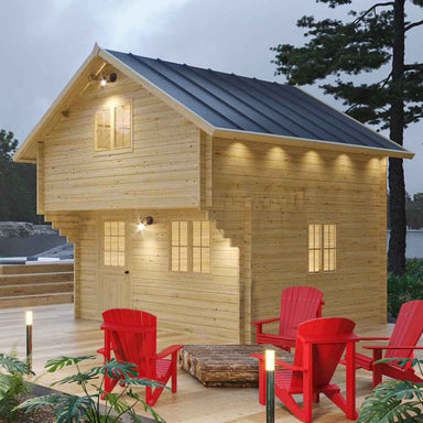 Lit by warm exterior lights, the Sawmill Structures The Jumbo Grand Loft 130 SD Garden Room sits on a wooden patio surrounded by red chairs and trees