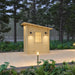 Straight front view of the Loo Loo House featuring a warm-lit glazed wooden door, matching black wall sconces, a sloped roof, and pathway lighting among forest greenery.