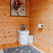 Loo Loo House bathroom interior with a white toilet beneath floral artwork, a small framed sign, wall-mounted toilet paper, a white trash bin, and a patterned fringed rug.