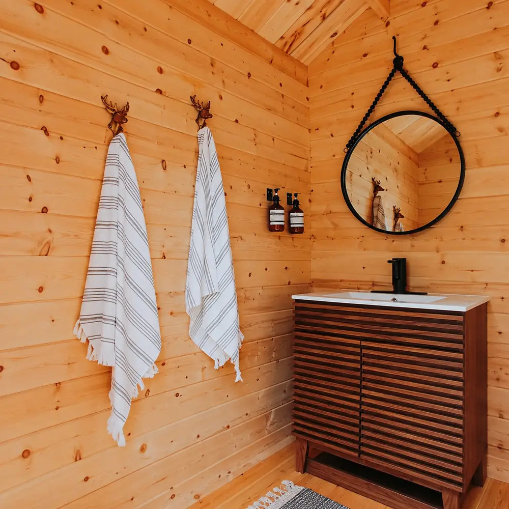 Loo Loo House bathroom interior with striped towels on antler hooks, amber soap dispensers, a round black-framed mirror, black faucet, and slatted wooden vanity.
