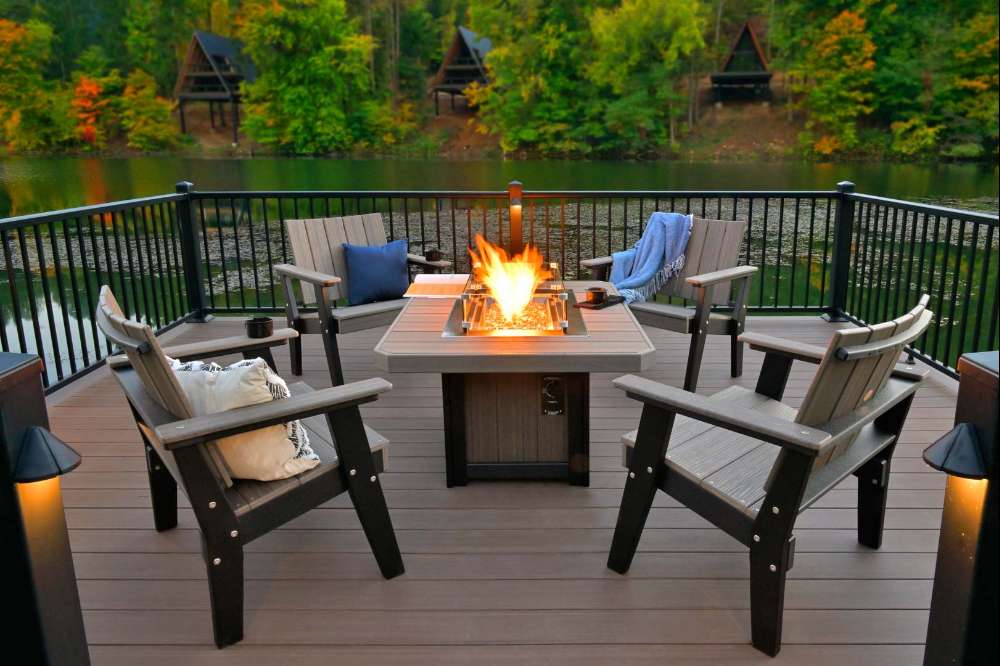 LuxCraft Hearthside Furniture Set surrounds a glowing fire pit on a deck overlooking a quiet lake and wooded cabins