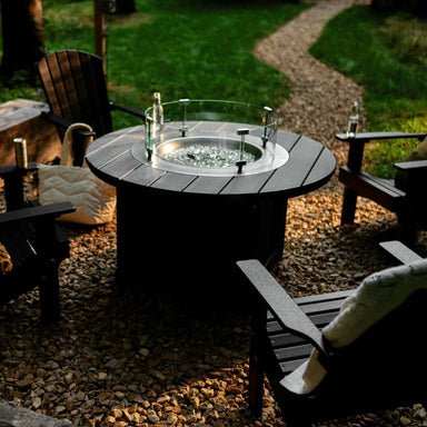 LuxCraft Lumin Fire Pit 46 Round Furniture Set features a round fire pit table with a glass wind guard, surrounded by Adirondack chairs on a gravel patio