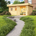 A vibrant garden scene showcases the GardenHouse24 USA Miriam 44B Classic Garden Room with open double doors revealing a cozy interior filled with framed artwork, greenery, and a soft sofa