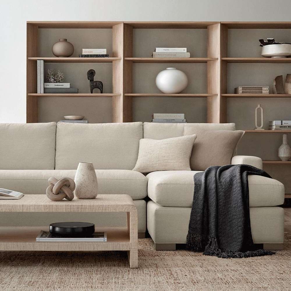 Modern living room uses Crate & Barrel Terrazza 114" Natural Solid Oak Wood 5-Shelf Bookcases, Set of 3 as a stylish backdrop behind a cream sectional sofa