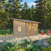 Angled view of the Multi Cube Versa 100 cabin with a slanted roof, wooden door, two glowing windows, deck, and forest landscaping.