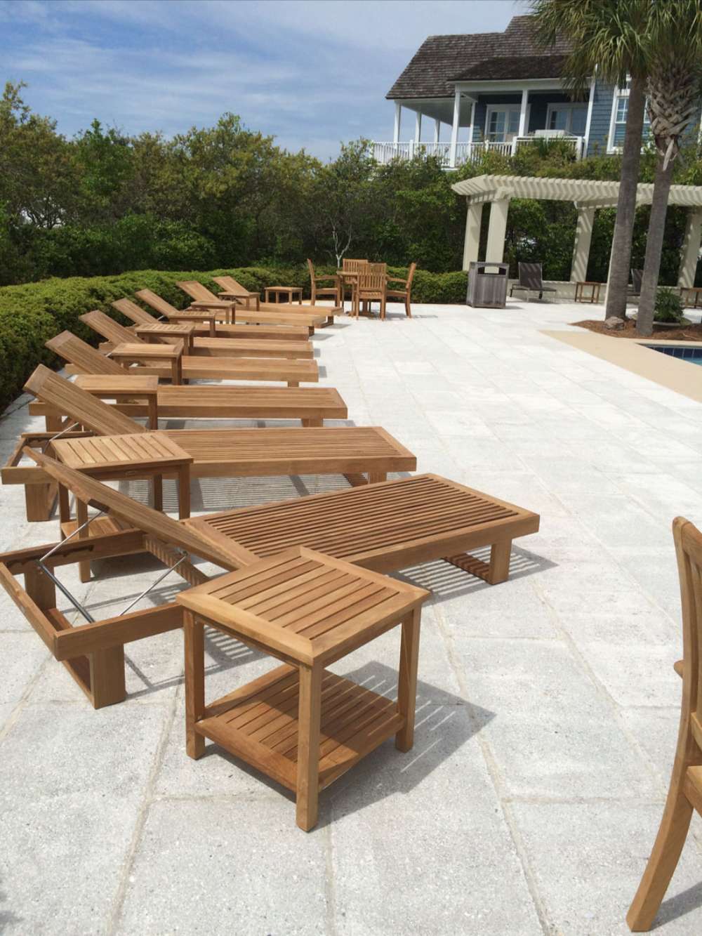 Multiple Westminster Teak Horizon Chaise Lounger chairs are arranged neatly around a spacious pool deck with palm trees and a house in the background