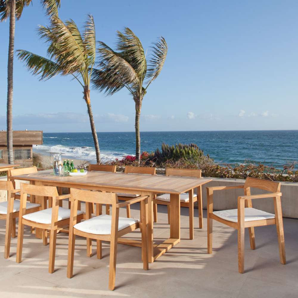 Multiple Westminster Teak Horizon Teak Dining Armchairs surround a large outdoor table on a patio overlooking the ocean, creating a relaxed dining setting