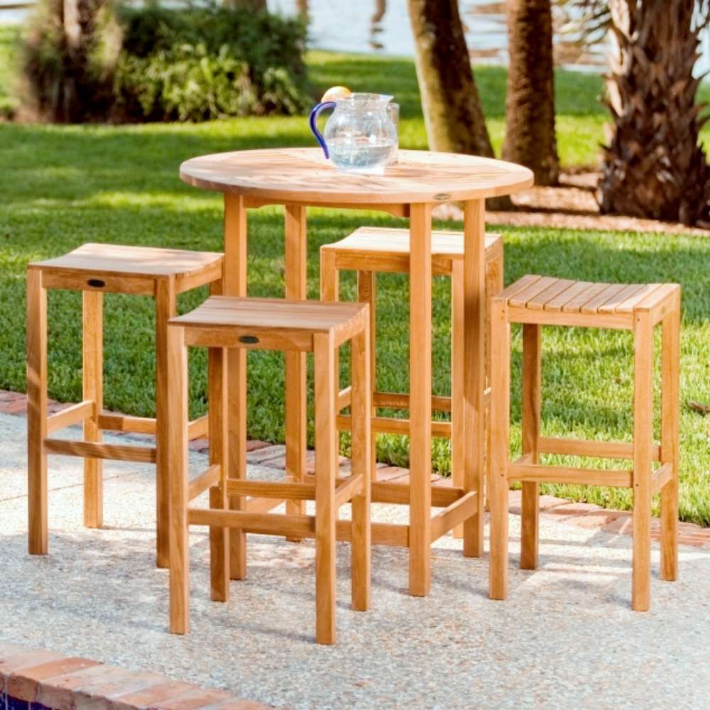 Multiple stools, including Westminster Teak Somerset Backless Bar Stool, surround a tall round outdoor table on a stone patio with trees and grass nearby