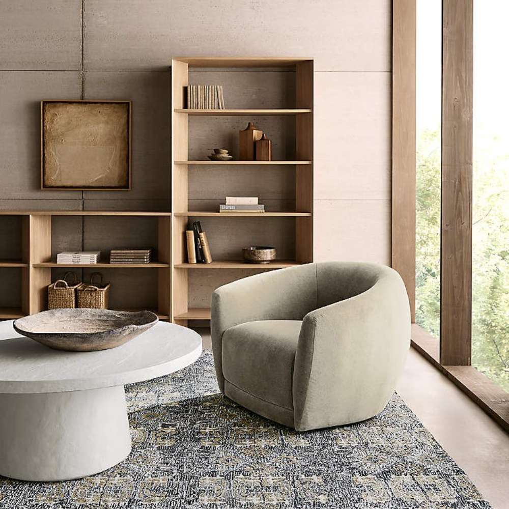 Next to a modern lounge chair and round coffee table, Crate & Barrel Terrazza 38" Natural Solid Oak Wood 5-Shelf Storage Bookshelf provides open display space for books and decor
