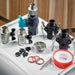 On a white surface, the valves, black hose connectors, drain grate, O-rings and thread seal tape for Golden Designs Dynamic Cold Therapy Cuboid 304 Nano-Coated Stainless Steel Cold Plunge Tub are arranged together