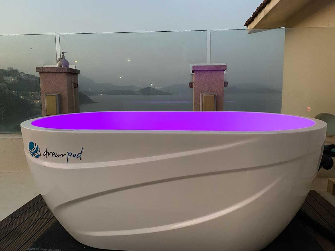 On an open balcony, a Dreampod Ice Bath with Chiller glows with purple lighting against a misty mountain and water view