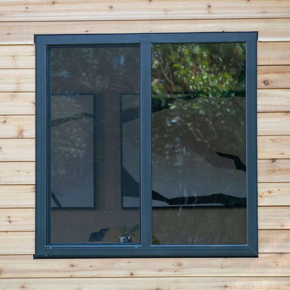 Outdoor Living Today 12×8 Saltbox Studio Outdoor Office is displayed in a close-up of a large window with dark trim reflecting trees and outdoor surroundings