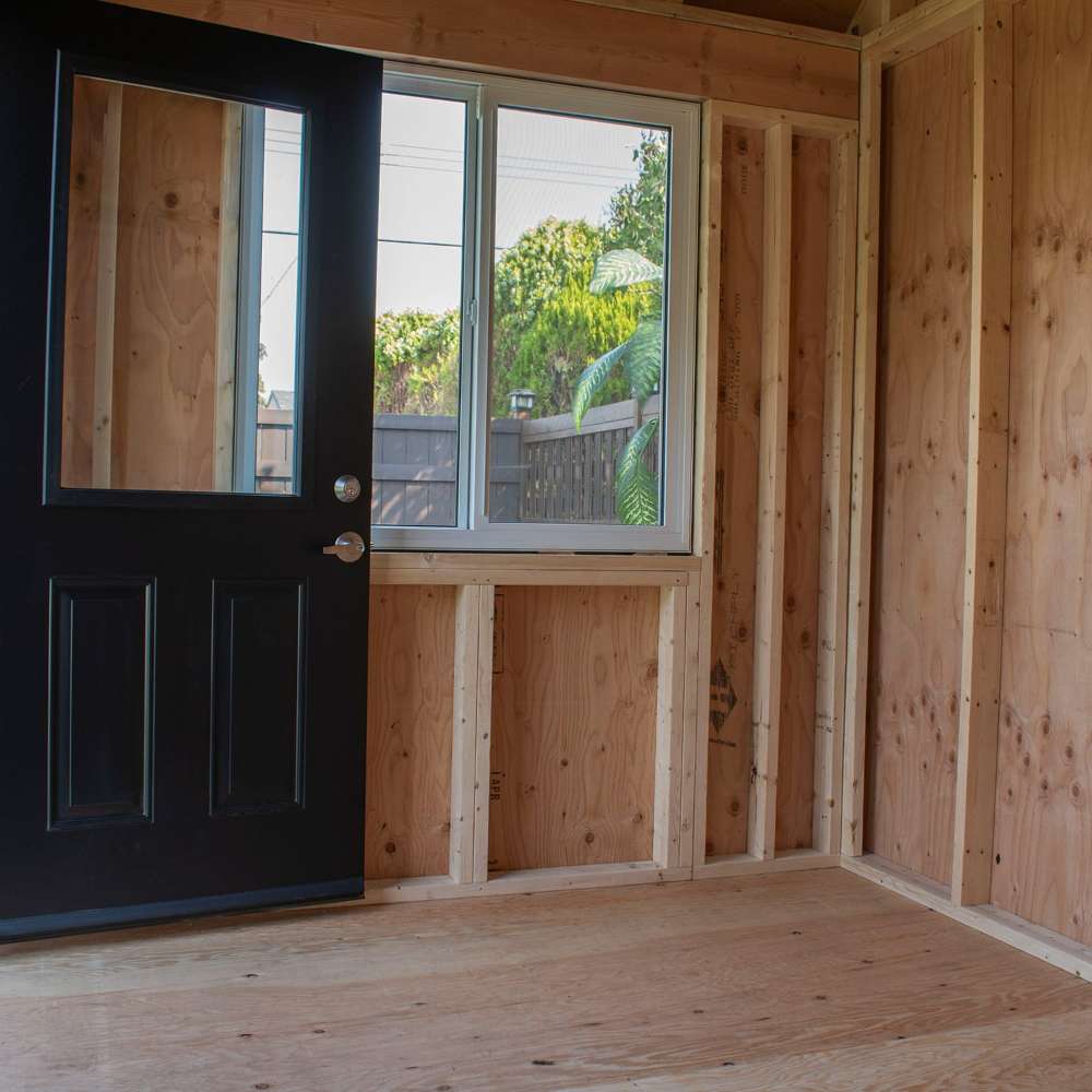 Outdoor Living Today 12×8 Saltbox Studio Outdoor Office is displayed inside with unfinished wooden walls, a black door, and a window letting in natural light