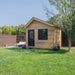 Outdoor Living Today 12×8 Saltbox Studio Outdoor Office is shown as a cedar shed placed on a green lawn with a black door, window, and surrounding trees in a backyard