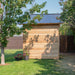 Outdoor Living Today 12×8 Saltbox Studio Outdoor Office is shown from the back as a cedar wall with a sloped metal roof surrounded by grass, trees, and potted flowers