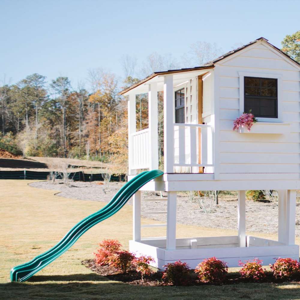 Outdoor Living Today 6x6 Little Cedar Playhouse & Sandbox features a white raised playhouse with a long green slide in a spacious yard