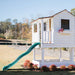 Outdoor Living Today 6x6 Little Cedar Playhouse & Sandbox features a white raised playhouse with a long green slide in a spacious yard