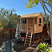 Outdoor Living Today 6x6 Little Cedar Playhouse & Sandbox includes a natural cedar playhouse with a porch, ladder access, and fenced backyard setting