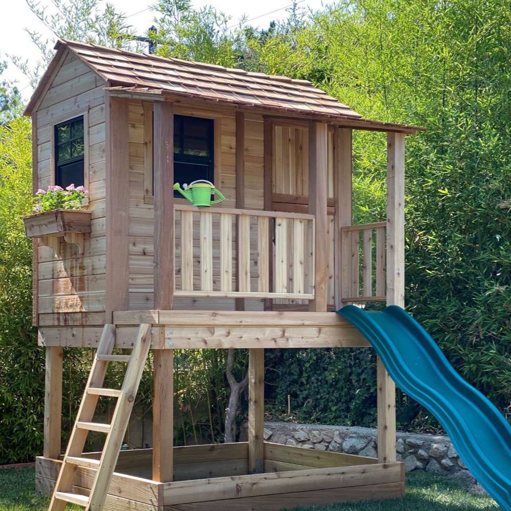 Outdoor Living Today 6x6 Little Cedar Playhouse & Sandbox includes an elevated wooden playhouse with a ladder, green slide, and sandbox space underneath