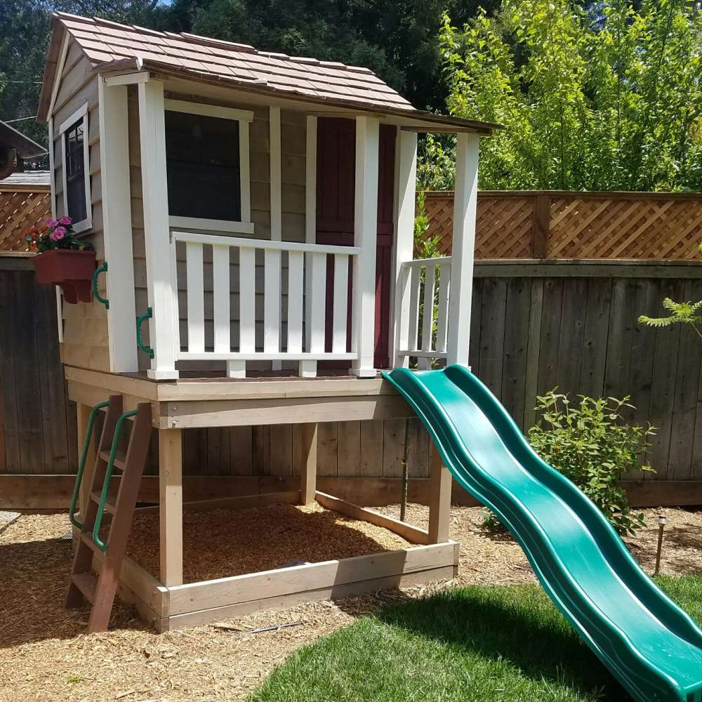 Outdoor Living Today 6x6 Little Cedar Playhouse & Sandbox includes white porch rails, side handles, and a long green slide in a backyard