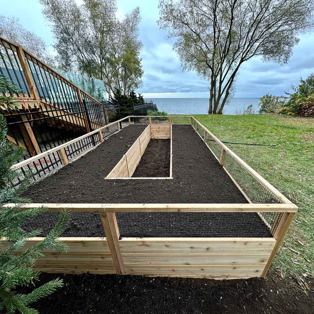 Outdoor Living Today 8×16 Raised Garden Bed sits filled with soil on a grassy lawn with a view of a lake and trees in the background