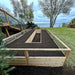 Outdoor Living Today 8×16 Raised Garden Bed sits filled with soil on a grassy lawn with a view of a lake and trees in the background