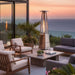 Outdoor Order PrismQuad Tower Heater glows on an ocean-view terrace with lounge chairs and a stone coffee table at sunset