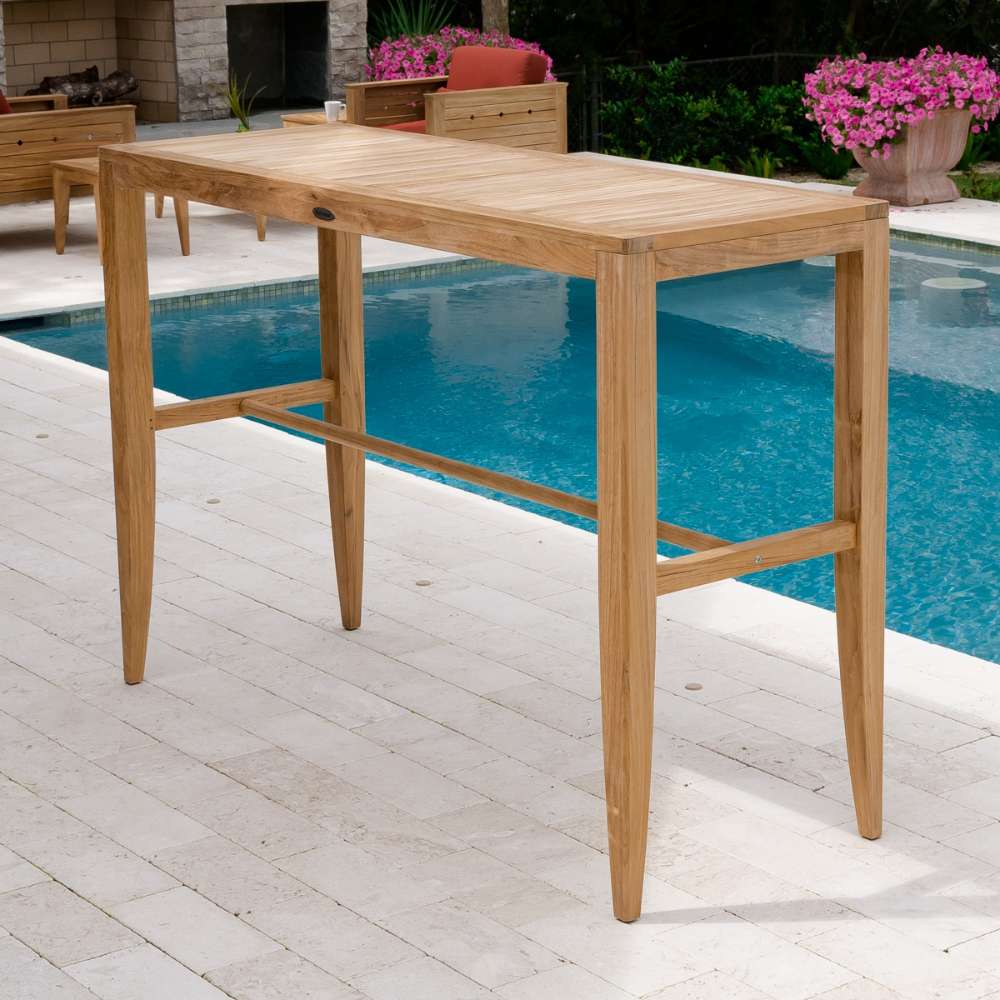 Outdoor teak bar table on a stone patio beside a pool with plants and a fireplace, styled from Westminster Teak Laguna 5pc Teak High Bar Set