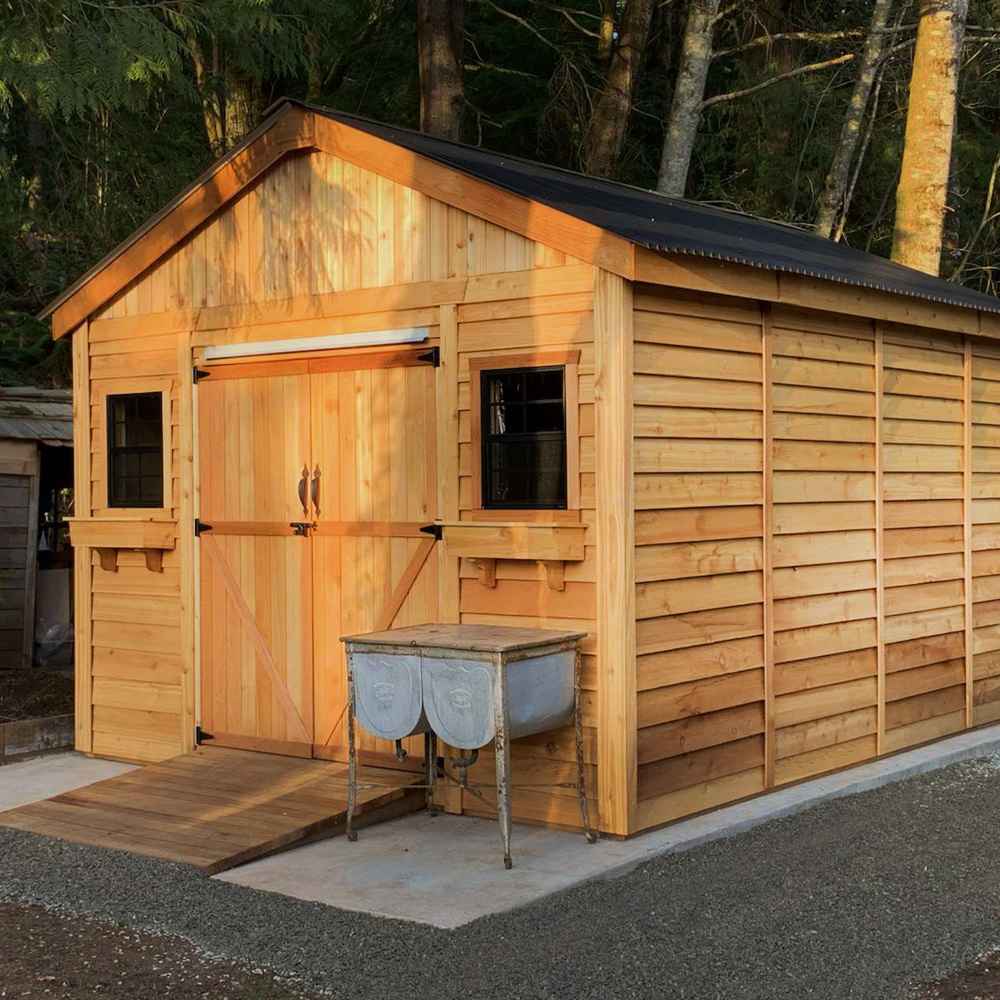 Outdoor Living Today 12×16 Double Door Space Master Storage Shed is a cedar structure with barn-style doors, black-trimmed windows, and a metal roof, sitting on a gravel foundation