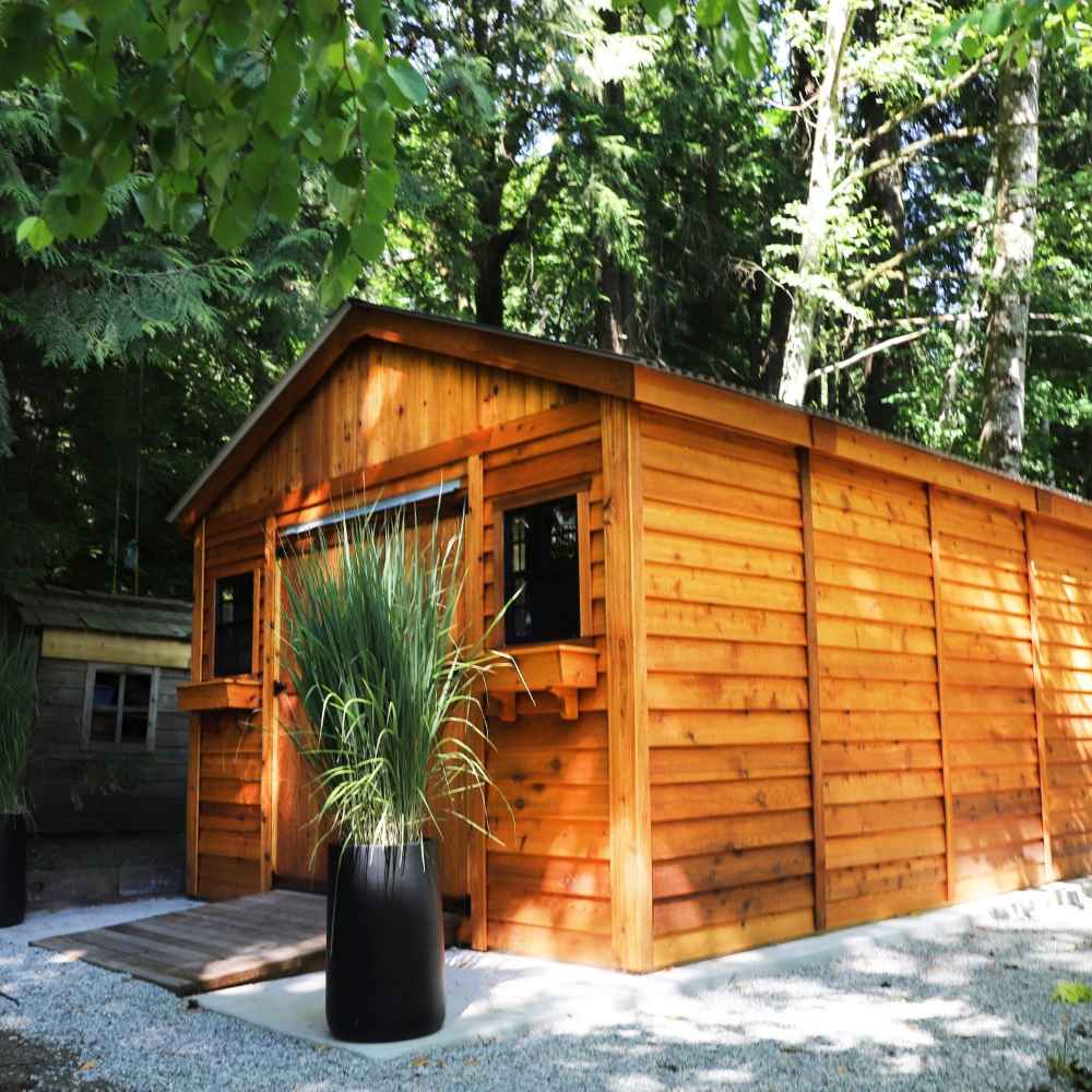 Outdoor Living Today 12×16 Double Door Space Master Storage Shed sits beautifully among tall trees, with potted plants flanking its entrance