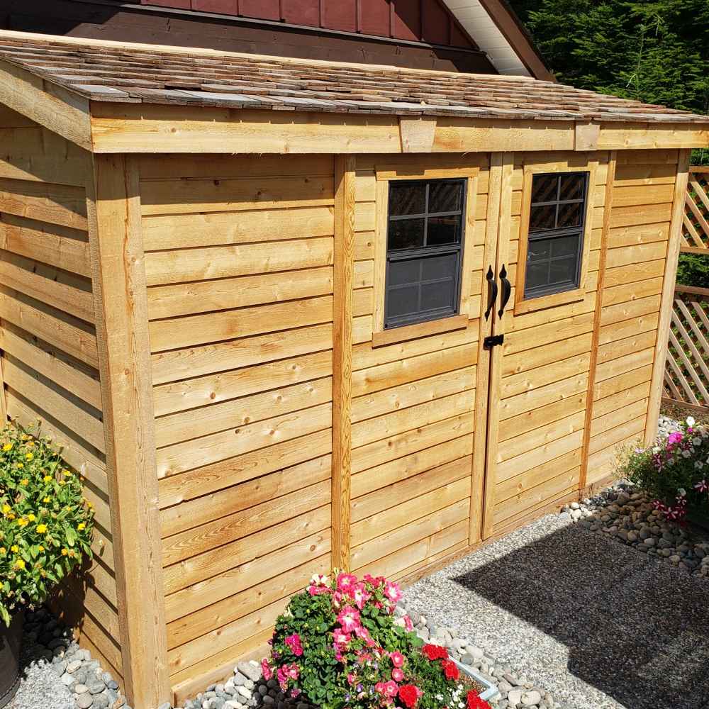 Outdoor Living Today 12×4 with Sliding Doors SpaceSaver Storage Shed is beautifully placed in a garden with blooming flowers surrounding its natural cedar exterior