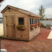Outdoor Living Today 8×12 Sunshed Garden Shed sits beside a waterfront on a red brick patio, with flower boxes lining its row of windows