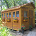 Outdoor Living Today 8×12 Sunshed Garden Shed sits outside in a leafy garden, decorated with red flowers in window boxes along its side