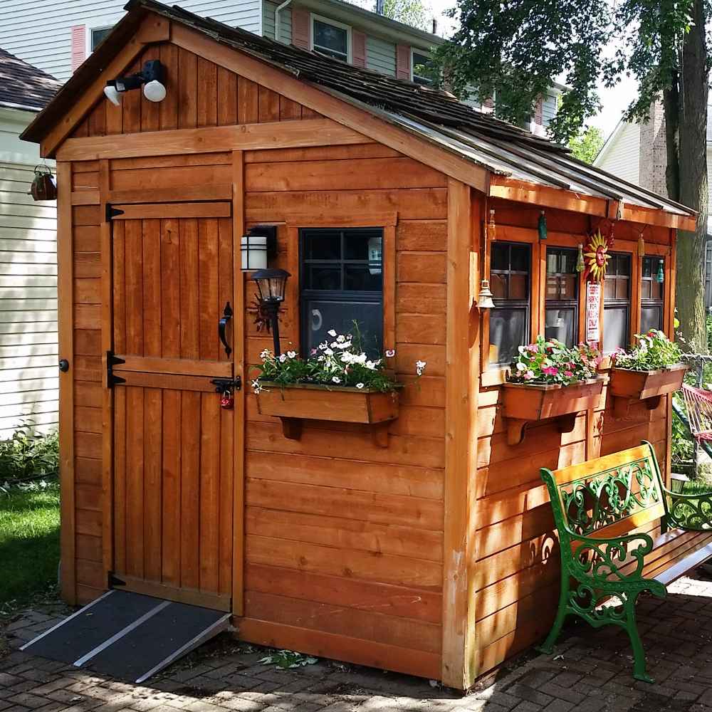 Outdoor Living Today 8×8 Sunshed Garden Shed is placed beside a house with flower-filled boxes, garden lighting, and a green bench in front
