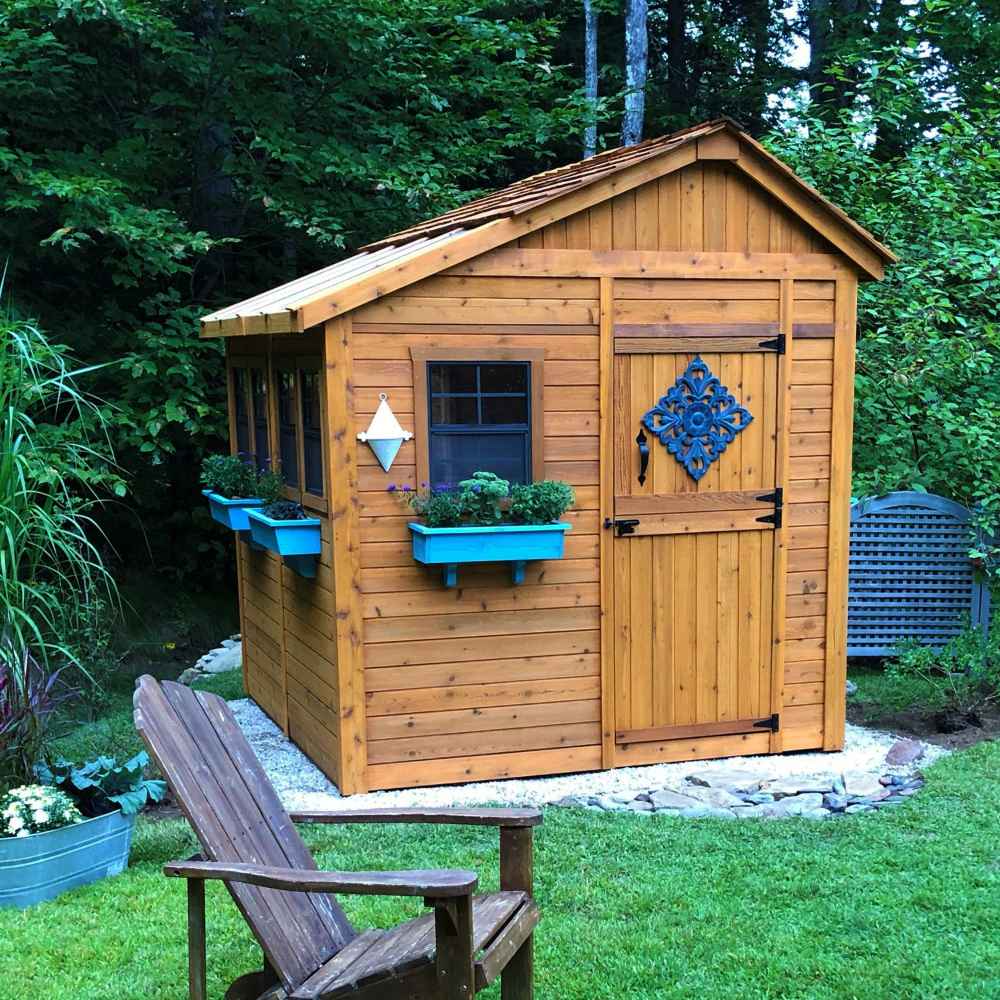 Outdoor Living Today 8×8 Sunshed Garden Shed sits in a wooded yard with blue flower boxes and a decorative design on the door