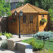 Outdoor Living Today 9×9 Penthouse Garden Shed glows warmly in the sunlight, framed by flowers and a decorative lamppost
