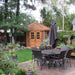 Outdoor Living Today 9×9 Penthouse Garden Shed sits at the back of a lush backyard, framed by patio furniture and trees