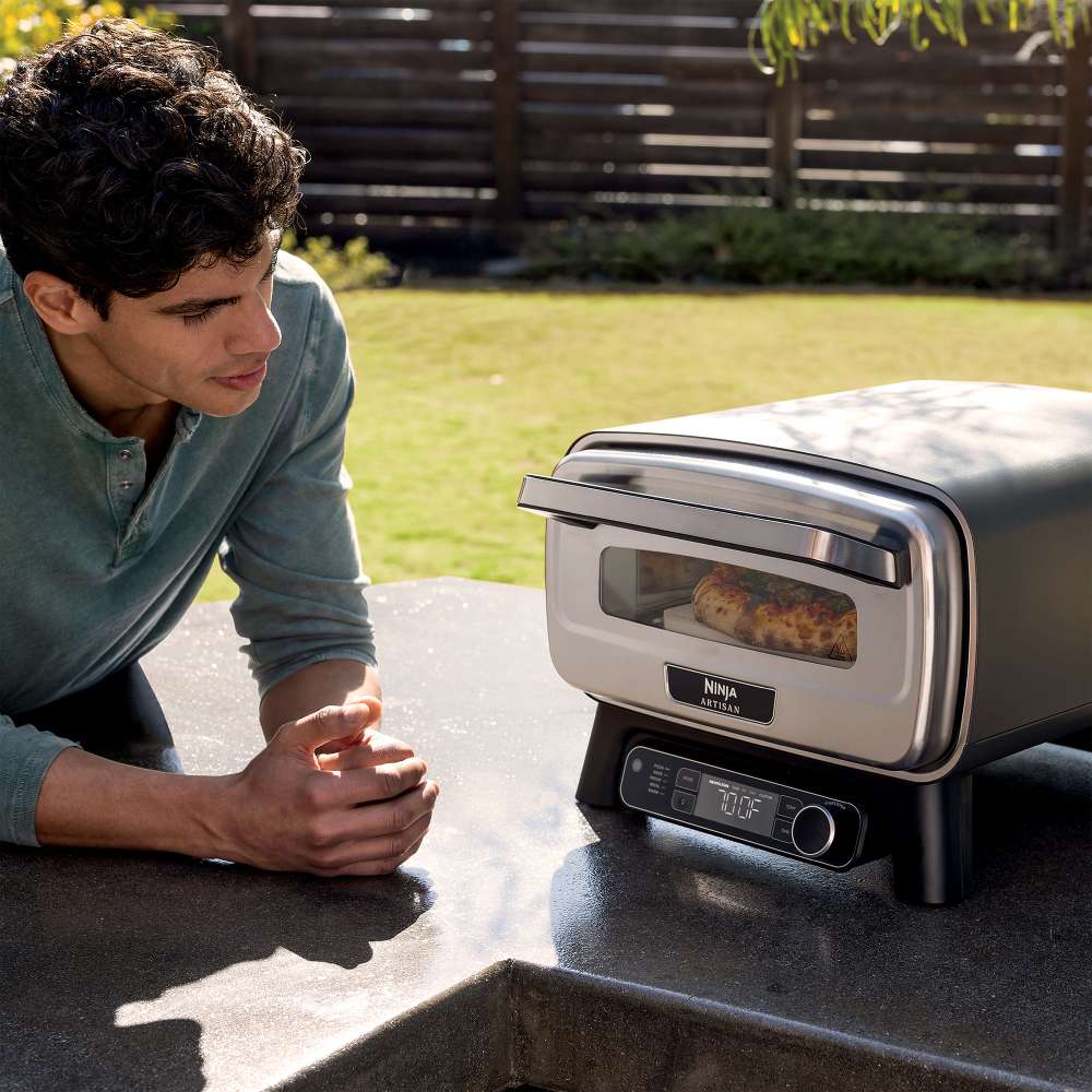 Outdoors, a person watches food cook through the front window of the Ninja Artisan 5-in-1 Portable Electric Pizza & Outdoor Oven