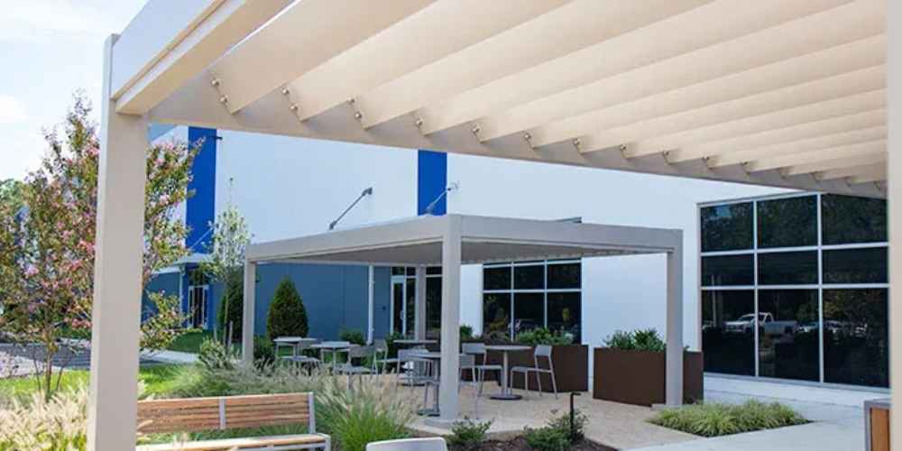 Outside a modern commercial building, the Structureworks Stratus Aluminum Pergola provides shaded seating and dining space amid landscaping with planters and benches