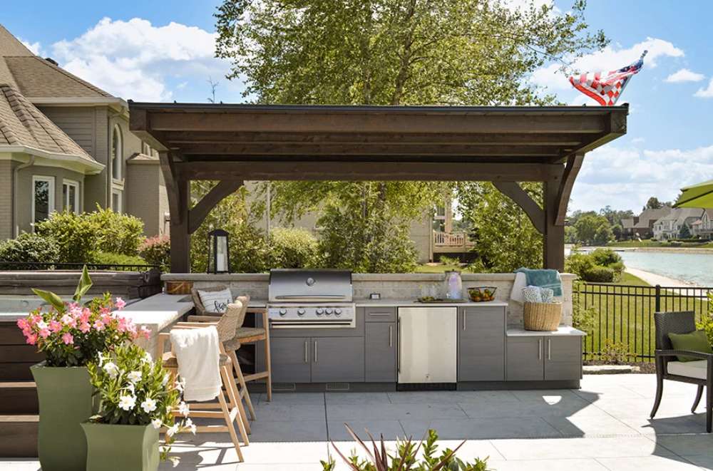 Overlooking a peaceful lake, the Brookside Timber Frame Ledgefield Pavilion provides shaded comfort for a sleek, modern outdoor kitchen with premium appliances