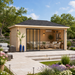 Pavilion ALU 44D Freedom Room fully furnished with cushioned seating, decorative pillows, lighting, and tables, set in a landscaped backyard garden with green grass, flowers, trees, and bright daytime surroundings.