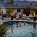 People swim in a backyard pool as two Outdoor Order Axis Collapsible Heater units glow beside the loungers at dusk