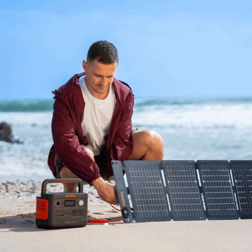 Person connects a foldable solar panel to Jackery Explorer 300 v2 Portable Power Station on a sandy beach for solar charging