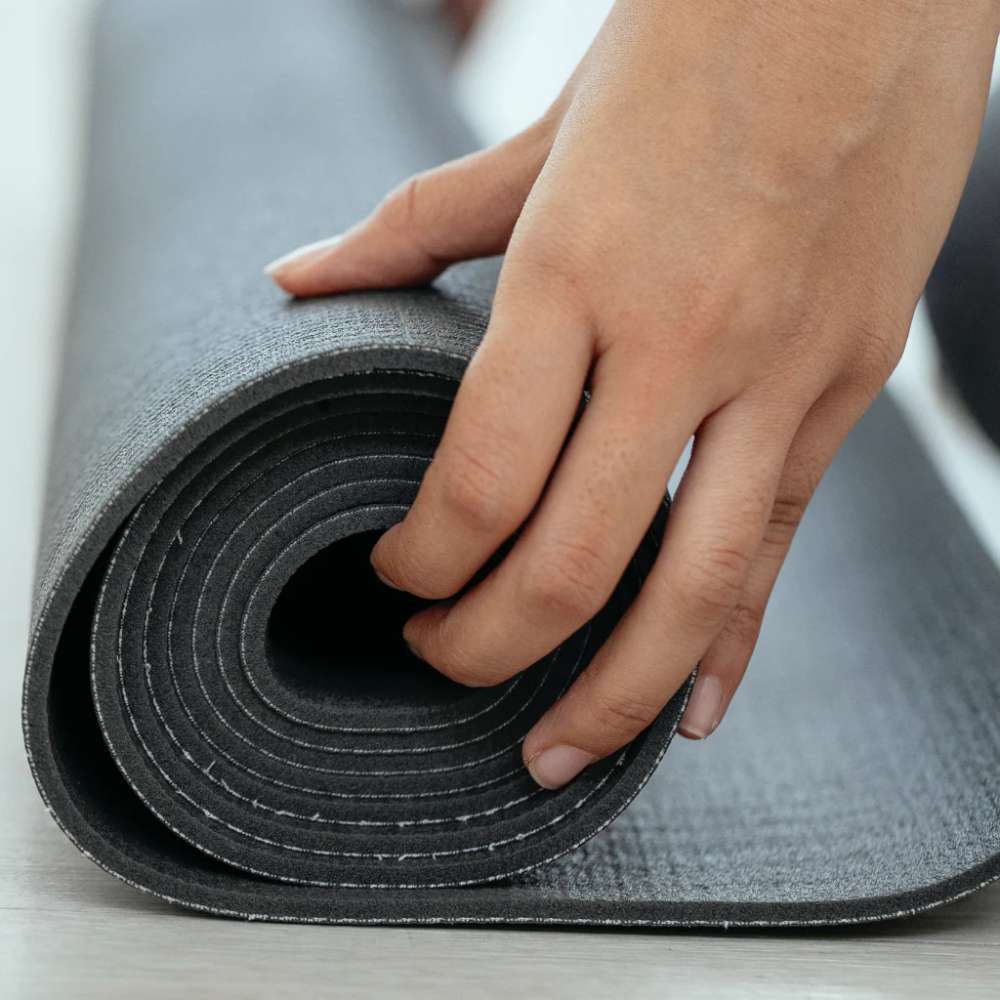 Person’s hand rolls up a thick black Dreampod Anti-Vibration Mat with a textured surface designed to reduce noise and vibration on hard floors